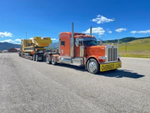 Bowline Logistics red semi-truck hauling oversized Keestrack machinery on a flatbed trailer under clear blue skies in the Alberta foothills