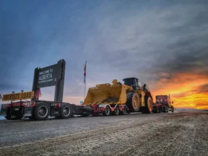 Bowline Logistics hauling a large tracked bulldozer on a red flatbed trailer through rural Alberta surrounded by autumn-coloured trees