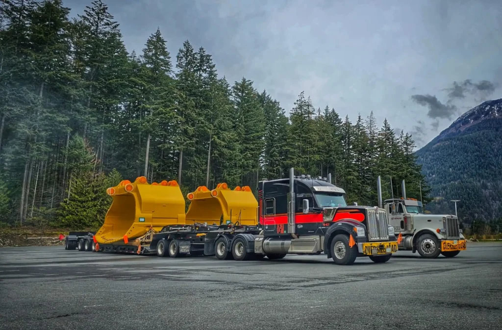 Two Bowline Logistics semi-trucks with "Oversize Load" banners are parked on a mountain pass, hauling three massive yellow excavator buckets against a backdrop of tall evergreen trees and misty mountain peaks Two Bowline Logistics semi-trucks with "Oversize Load" banners are parked on a mountain pass, hauling three massive yellow excavator buckets against a backdrop of tall evergreen trees and misty mountain peaks