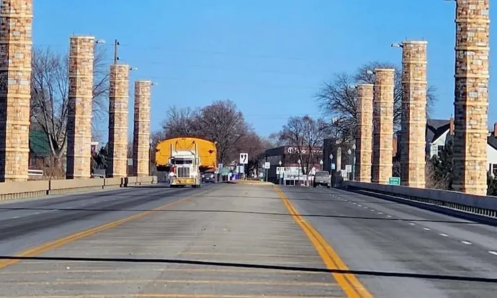Just In Time Transport Oversized yellow load transported by a Bowline Logistics truck down a city street lined with tall stone pillars under clear skies