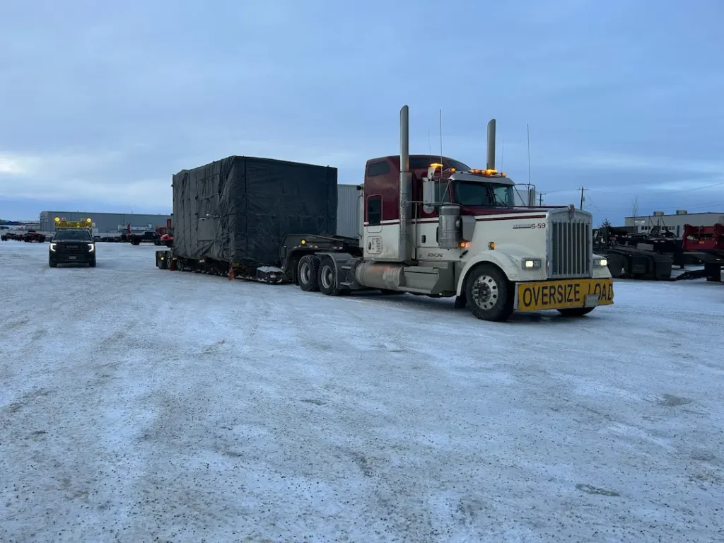 Bowline Logistics semi-truck hauling a large black-wrapped project cargo load on a flatbed trailer across a snowy yard Bowline Logistics semi-truck hauling a large black-wrapped project cargo load on a flatbed trailer across a snowy yard